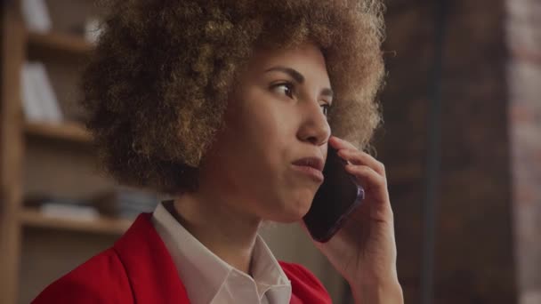 穿着红色夹克的非裔美国女商人在舒适的阁楼办公室里打电话聊天时面带微笑 — 图库视频影像
