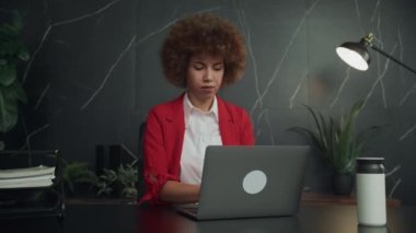 Kendine güvenen, kırmızı ceketli profesyonel bir kadın, şık bir ofis ortamında dizüstü bilgisayarı inceliyor.