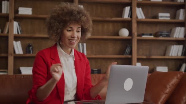一个兴奋的女人 卷曲的头发在笔记本电脑前欢呼 穿着红色夹克 躺在舒适的办公室里 — 图库视频影像