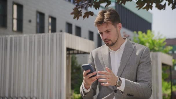 灰色のスーツを着た若いビジネスマンは 都市の建物の外で彼の携帯電話にメッセージを読んで心配しているように見えます — ストック動画