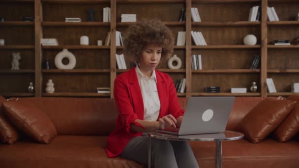 在时尚的办公室休息室使用笔记本电脑 集中注意力的年轻女性 一头卷曲的头发 穿着红色夹克 — 图库视频影像