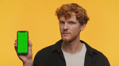 Kızıl saçlı genç adam, sarı arka planda krom tuşlu yeşil ekranlı akıllı telefonu sunuyor. Hiç bir hareket yapmadan başını sallıyor.