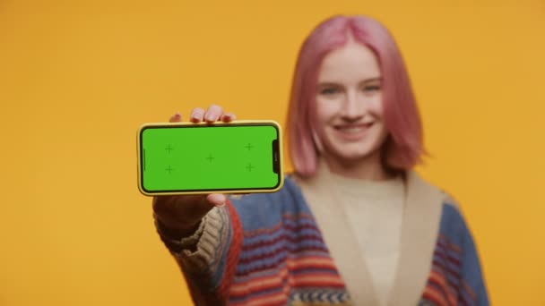 一位快乐的年轻女子 一头生机勃勃的粉色头发 展示了一款带有绿色屏幕的智能手机 非常适合在黄色背景下演示应用程序 — 图库视频影像