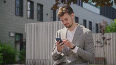 İyi giyimli, şık, gri takım elbiseli bir adam odaklanmış bir yüzüyle akıllı bir telefon kullanıyor arka planda bir şehir binası olan açık havada.