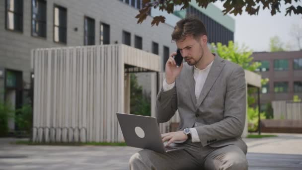 一位专心致志的创业者 穿着配套的灰色西装 一边在城市花园的手机上聊天 一边用笔记本电脑完成多项任务 — 图库视频影像