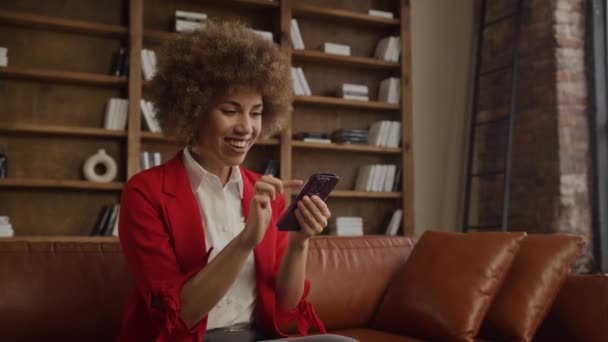 一位穿着红色西装的女商人坐在阁楼式的房间里 看着她的智能手机 高兴极了 — 图库视频影像