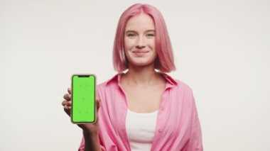 Pembe saçlı gülümseyen kadın beyaz arka planda yeşil ekranlı akıllı bir telefon tutuyor.