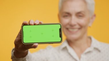 Gülümseyen yaşlı kadın yeşil ekranlı akıllı telefonu sunuyor. Krom tuşlu telefona odaklanıyor.