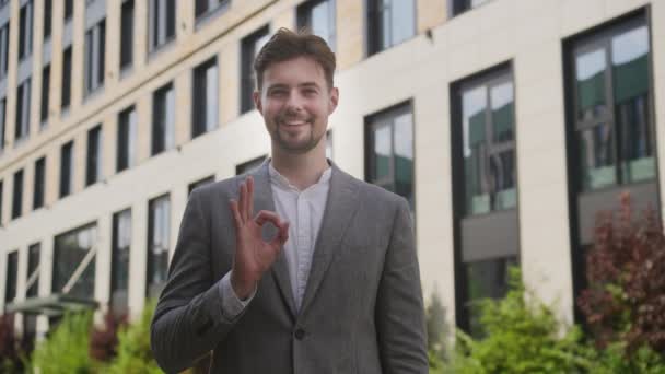 Okのジェスチャーを作るスマートなカジュアルなスーツで陽気な若いビジネスマン 近代的なオフィスビルの外で承認または成功を伝える — ストック動画