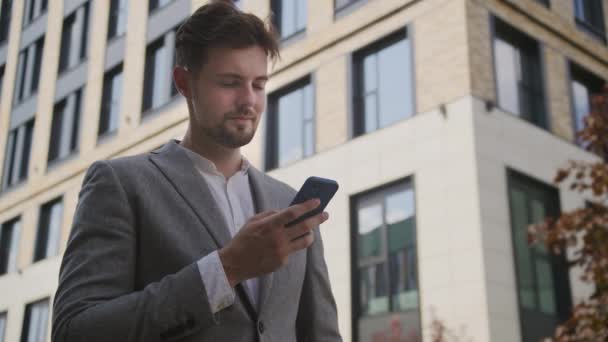 一位穿着灰色西装的专注的年轻商人在现代写字楼外使用智能手机 沉浸在科技之中 — 图库视频影像