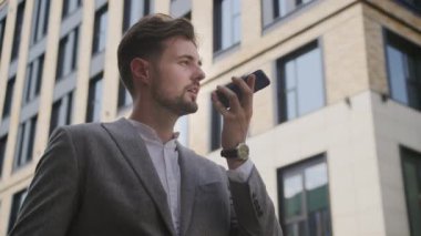Akıllı gri takım elbiseli odaklanmış genç bir adam bir ofis binasının dışındaki akıllı telefonundan sesli mesaj kaydediyor.