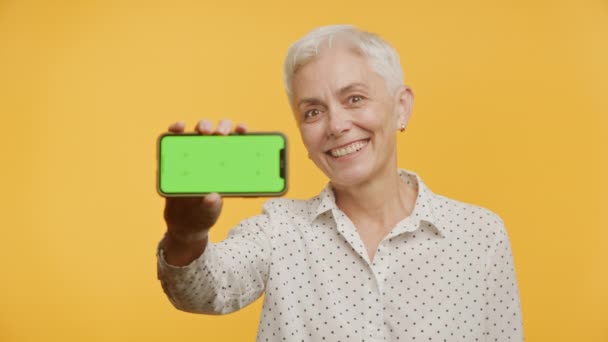 Uśmiechnięta Starsza Pani Prezentuje Telefon Zielonym Ekranem Odizolowany Żółtym Tle — Wideo stockowe