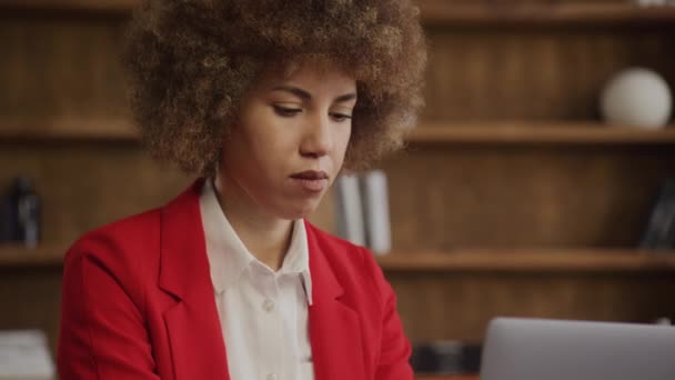 ラップトップ画面 オフィスの背景に焦点を当てた赤いジャケットのカーリーヘアの深刻な若い女性 — ストック動画