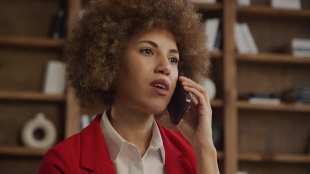 焦点を当てた表現 オフィスの本棚の背景を持つ携帯電話で話している赤い黒い髪の女性 — ストック動画