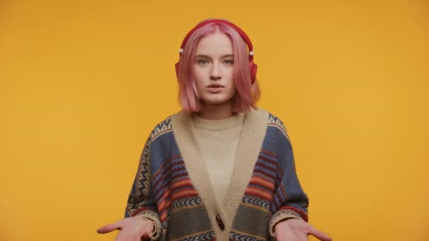 ピンクの髪とヘッドフォンの笑顔の女性は 喜んでビデオ通話で話し 黄色の背景に対して手を挙げます — ストック動画