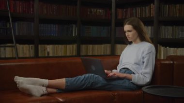 Dizüstü bilgisayarında çalışırken yorgun görünen genç bir kadın kütüphanede deri bir koltukta oturuyor.