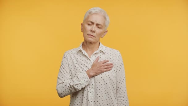 在黄色背景下表现出诚实的老年妇女 双手捂住心口 — 图库视频影像