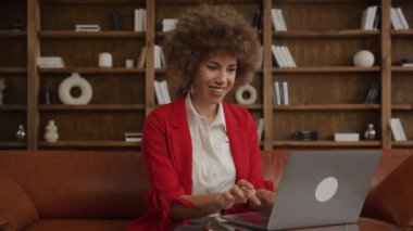 Kıvırcık saçlı, gülümseyen genç bir kadın bilgisayarla olumlu bir mesaj okuyor, kırmızı ceket giymiş, modern ofiste kapalı bir yerde.