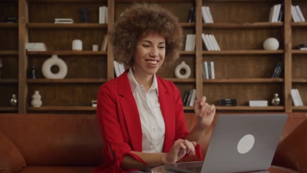 楽しい若い女性 彼女のラップトップ上のビデオ通話中に挨拶のジェスチャーで手を振る赤いスーツ — ストック動画