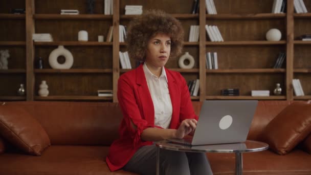 彼女のラップトップで作業しながら親指を与えるカーリーヘアと笑顔の若い女性は オフィスで赤いジャケットを着ています — ストック動画