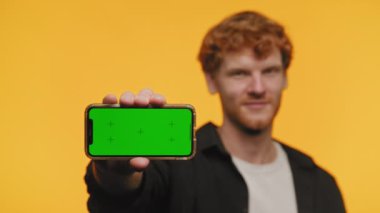 Kızıl saçlı genç adam yeşil ekranlı cep telefonunu canlı turuncu bir arkaplana karşı içerik değişikliğine hazır olarak sunuyor.