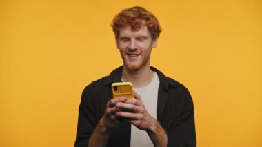 Neşeli kızıl saçlı genç adam cep telefonuyla mesaj atıyor, canlı sarı bir fon karşısında neşeli bir anın tadını çıkarıyor.
