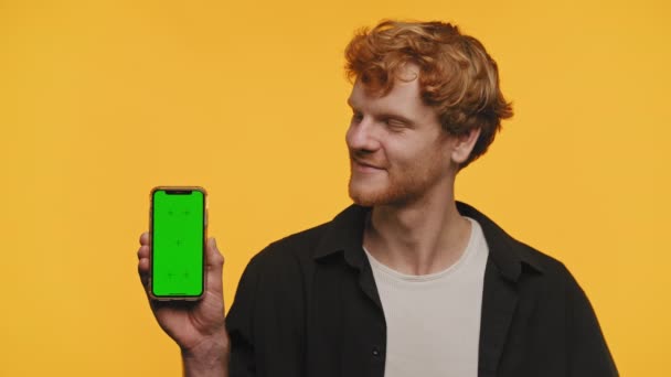 黄色の背景にクロマキー画面の携帯電話を提示するカジュアルな服装で陽気なカーリー髪の男 — ストック動画