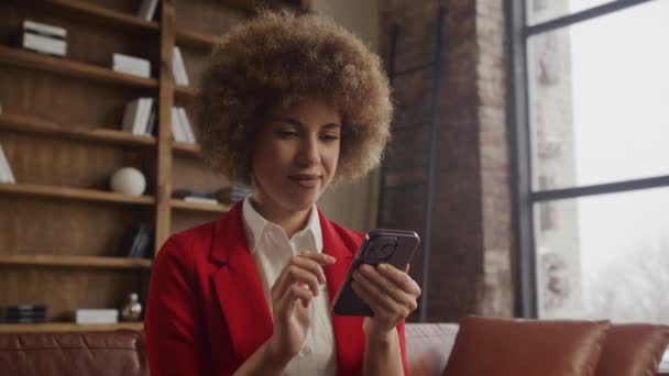 卷发女商人穿着红色西装 在舒适的阁楼工作间用她的智能手机 — 图库视频影像