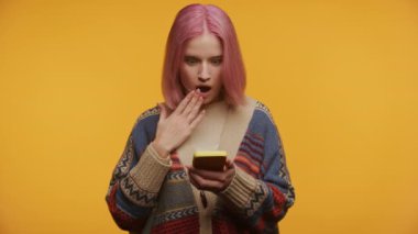 Pembe saçlı genç bir kadın sarı arka planda sarı bir telefona bakarken şok olmuş.