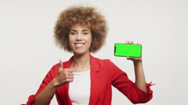 Kıvırcık saçlı neşeli kadın yeşil ekranlı telefonu gösteriyor, başparmağını kaldırıyor.