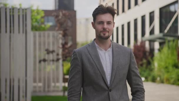バックグラウンドのモダンなオフィスビルで屋外に立っている灰色のスーツの陽気な若いビジネスマン — ストック動画