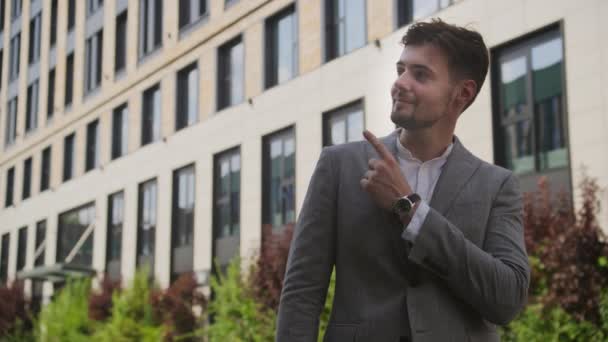 上を向くカジュアルなグレーのスーツで笑顔のビジネスマン オフィスの建物の背景を持つ緑豊かな庭に立って — ストック動画