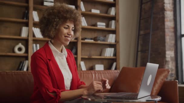 一位穿着红色西装的快乐的女商人在时尚的阁楼工作区用笔记本电脑进行视频通话时挥手致意 — 图库视频影像
