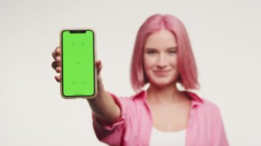 Pembe saçlı genç kadın, beyaz arka planda yeşil ekranlı akıllı telefonu sunuyor.