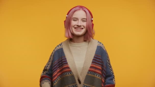 黄色の背景に対するビデオ通話でこんにちは振るピンクの髪とヘッドフォンを持つ笑顔の女性 — ストック動画