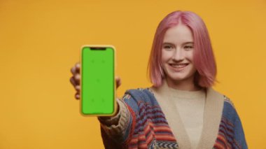 Pembe saçlı neşeli öğrenci sarı arkaplanda yeşil ekran renkli bir telefon sunuyor.