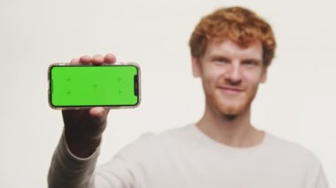 Elinde yeşil ekranlı, beyaz arka planda izole bir telefon tutan gülümseyen kızıl adam.