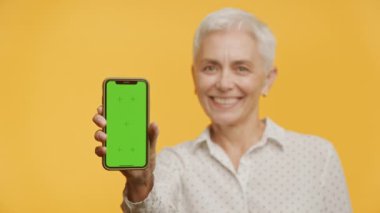 Neşeli olgun bir kadın yeşil ekranlı bir telefon gösteriyor. Özelleştirmeye hazır, sarı bir arka planda.