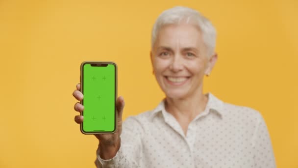 Весела Зріла Жінка Показує Телефон Зеленим Екраном Готовий Налаштування Жовтому — стокове відео