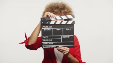 Beyaz arka planda film çekmeye hazır bir kadın el çırpma tahtasından gizlice bakıyor.