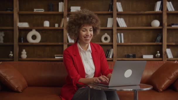 ラップトップ 快適なオフィスの設定を使用して赤いブレーザーでカーリーヘアの陽気な若い女性 — ストック動画