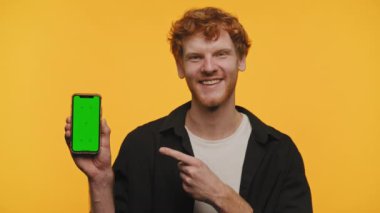 Sarı arka planda yeşil ekranlı akıllı bir telefonu işaret eden kıvırcık saçlı neşeli bir adam.