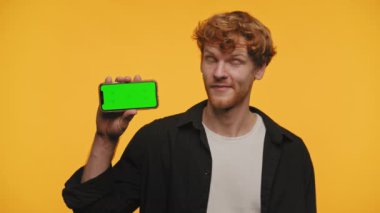 Kıvırcık saçlı hoşnutsuz adam yeşil ekranlı akıllı telefona baş parmak gösteriyor.