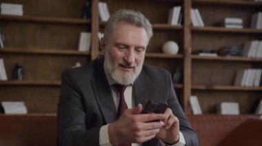 Gülümseyen olgun erkek yönetici rahat bir ofis ortamında telefonla mesajlaşıyor.