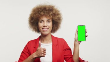 Neşeli kadın yeşil ekranlı akıllı telefonu gösterirken başparmağını kaldırıyor.