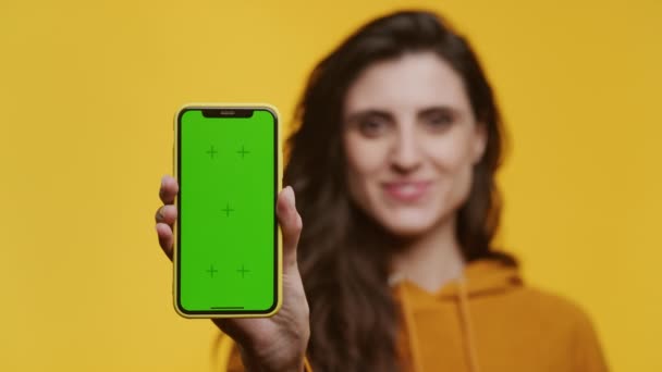 穿着橙色帽衫的微笑女人举着智能手机 绿色屏幕上的彩色键与黄色背景隔离 — 图库视频影像