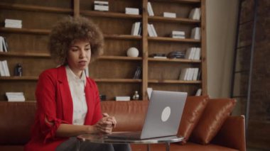 Kırmızı elbiseli kıvırcık saçlı bir kadın ofiste dizüstü bilgisayar kullanarak online toplantı yapıyor.