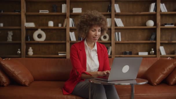 穿着红色西装的女人坐在时尚办公室的皮沙发上 一边在笔记本电脑上工作 一边笑 — 图库视频影像