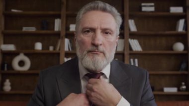 Sakallı, olgun bir adam. Ofis ortamında kravatını düzeltiyor.