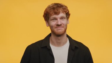 Kızıl saçlı, gülümseyen adam sarı arka planda çift parmak işareti yapıyor ve başını sallıyor.